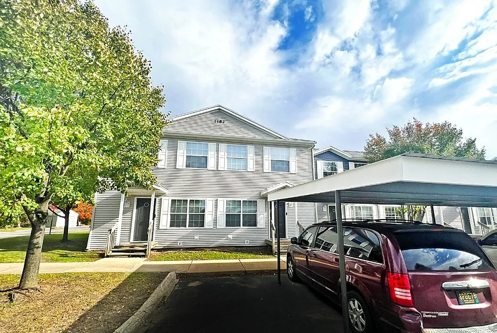 Charming suburban townhouse with two stories, large front windows, and a covered parking area, nestled in a serene neighborhood with mature trees.