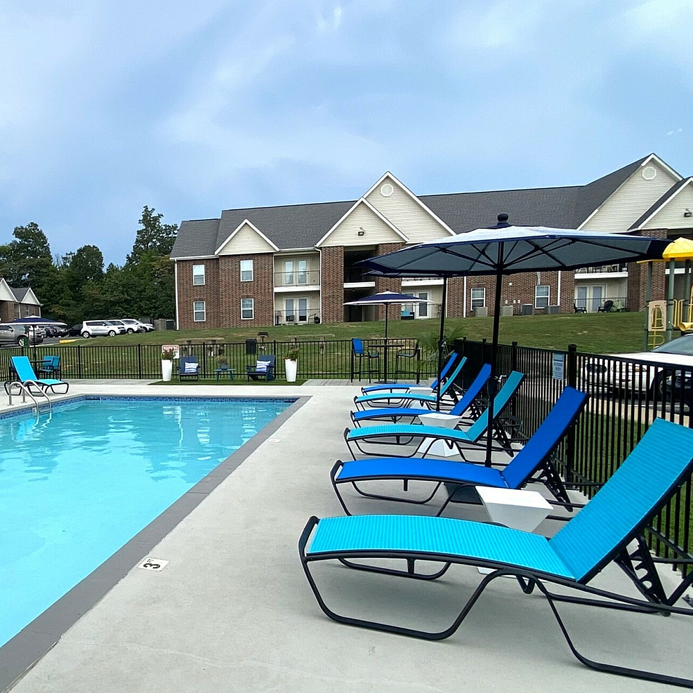 Inviting apartment complex pool area with vibrant blue loungers and umbrellas, surrounded by well-maintained buildings and a playground in the background.