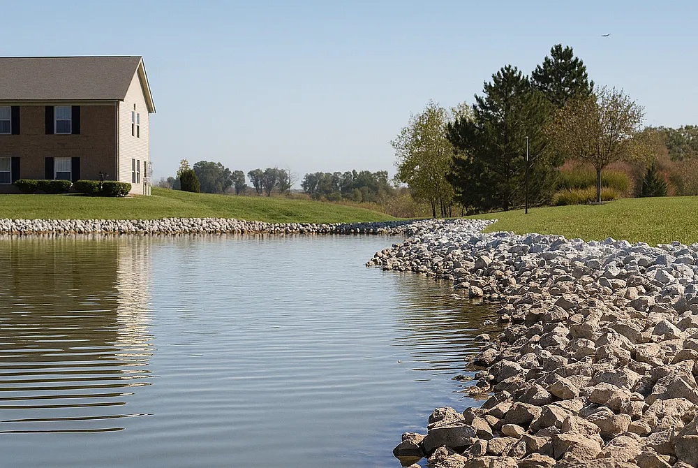 Serene riverside view with a charming house, lush greenery, and a tranquil stone-lined shoreline.