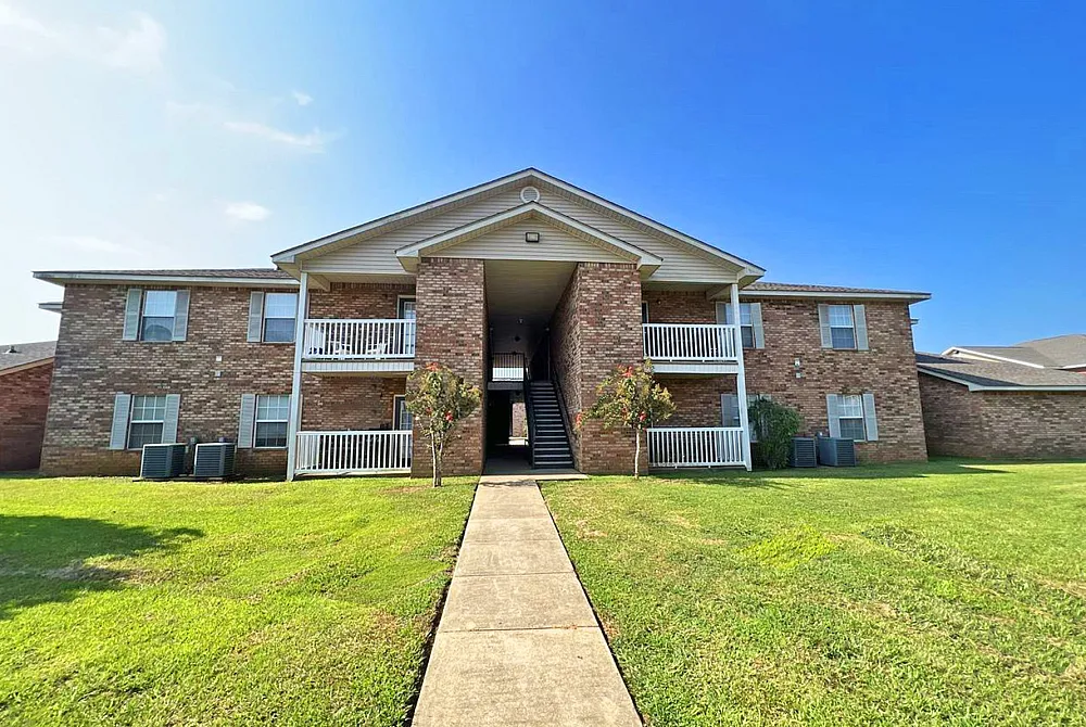 Charming brick apartment complex showcasing spacious balconies, lush green lawns, and a welcoming entrance under a clear blue sky.
