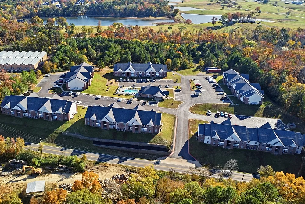 Scenic aerial view of a charming apartment complex surrounded by lush greenery and tranquil lakes, featuring ample parking and inviting outdoor spaces.