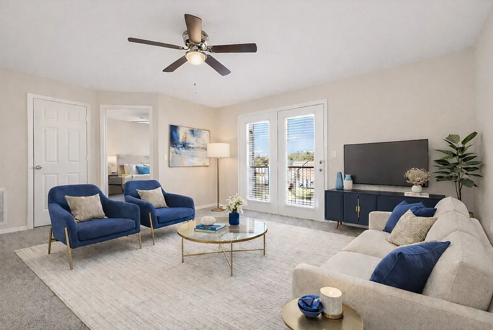 Inviting living room with modern decor, featuring blue accent chairs, a cozy beige sofa, and a glass coffee table under a stylish ceiling fan, bathed in natural light from large windows.