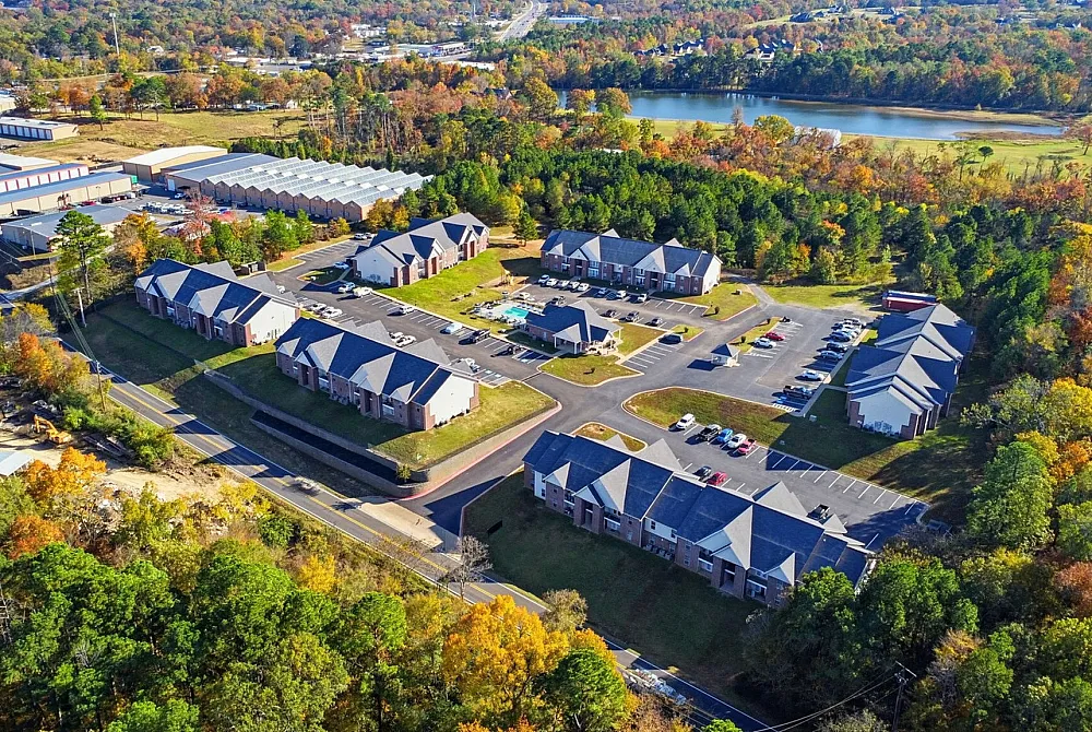 Scenic aerial view of tranquil apartment community surrounded by lush greenery and a serene lake, offering a peaceful living environment with spacious parking and modern amenities.