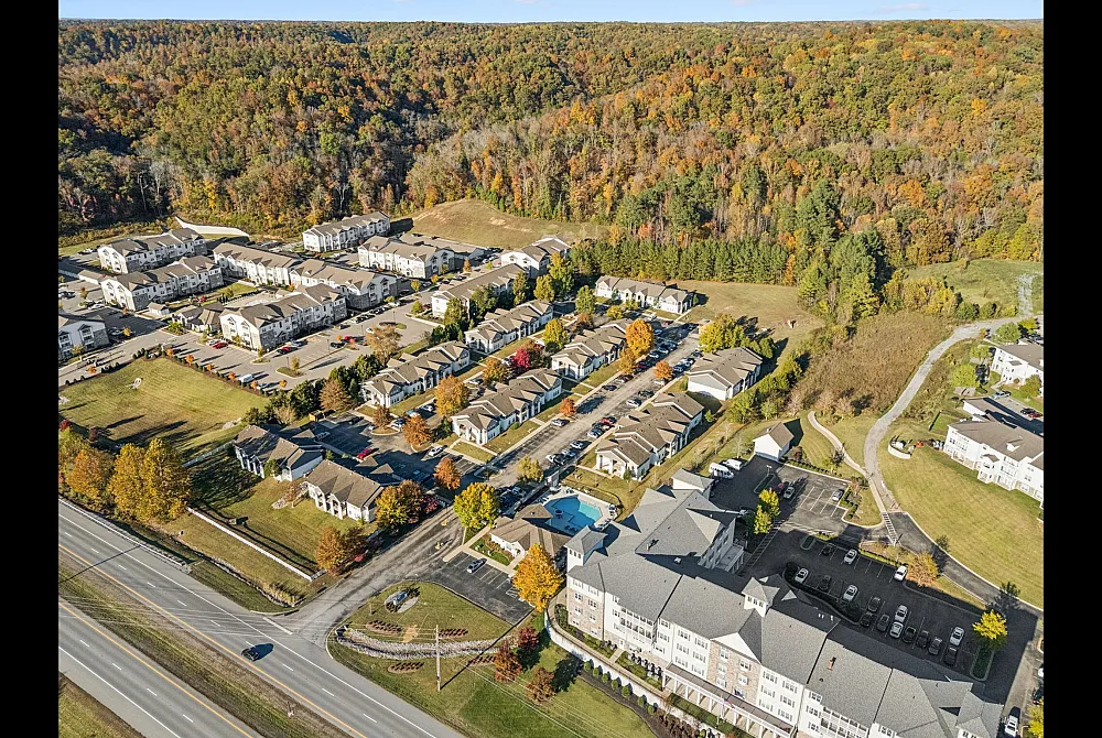 Scenic aerial view of a modern apartment complex nestled in lush greenery with vibrant autumn colors, showcasing ample parking, pristine roads, and a community pool.