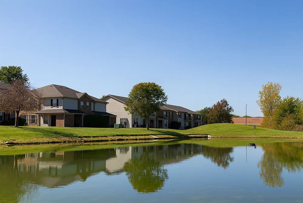 Picturesque residential area with modern houses beside a tranquil pond, surrounded by lush greenery and clear blue skies.