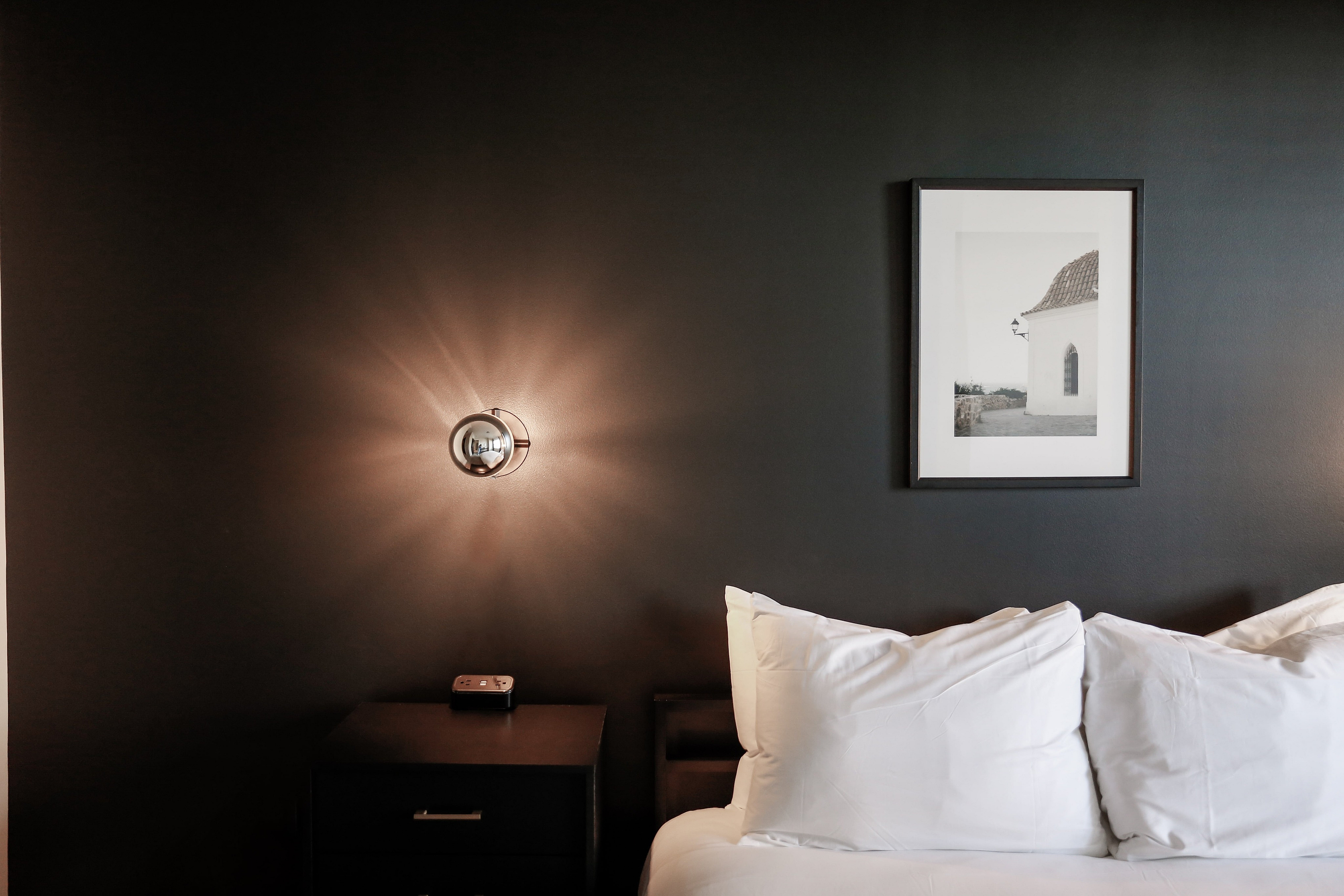 Elegant bedroom featuring a dark accent wall, modern wall sconce, and cozy white bedding with a framed artistic print.