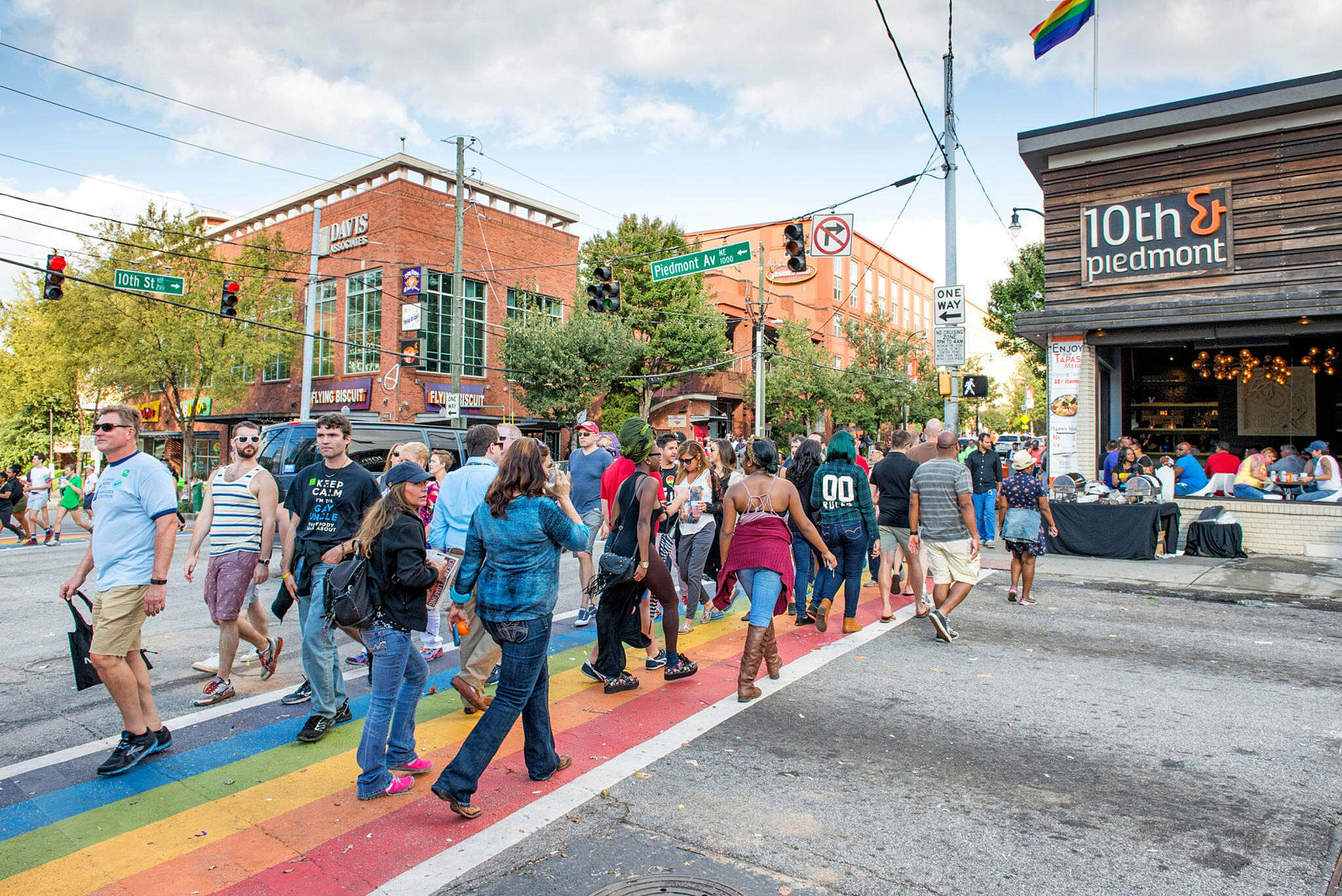 Vibrant urban street scene with a diverse crowd walking along a colorful rainbow crosswalk, surrounded by lively restaurants and shops.