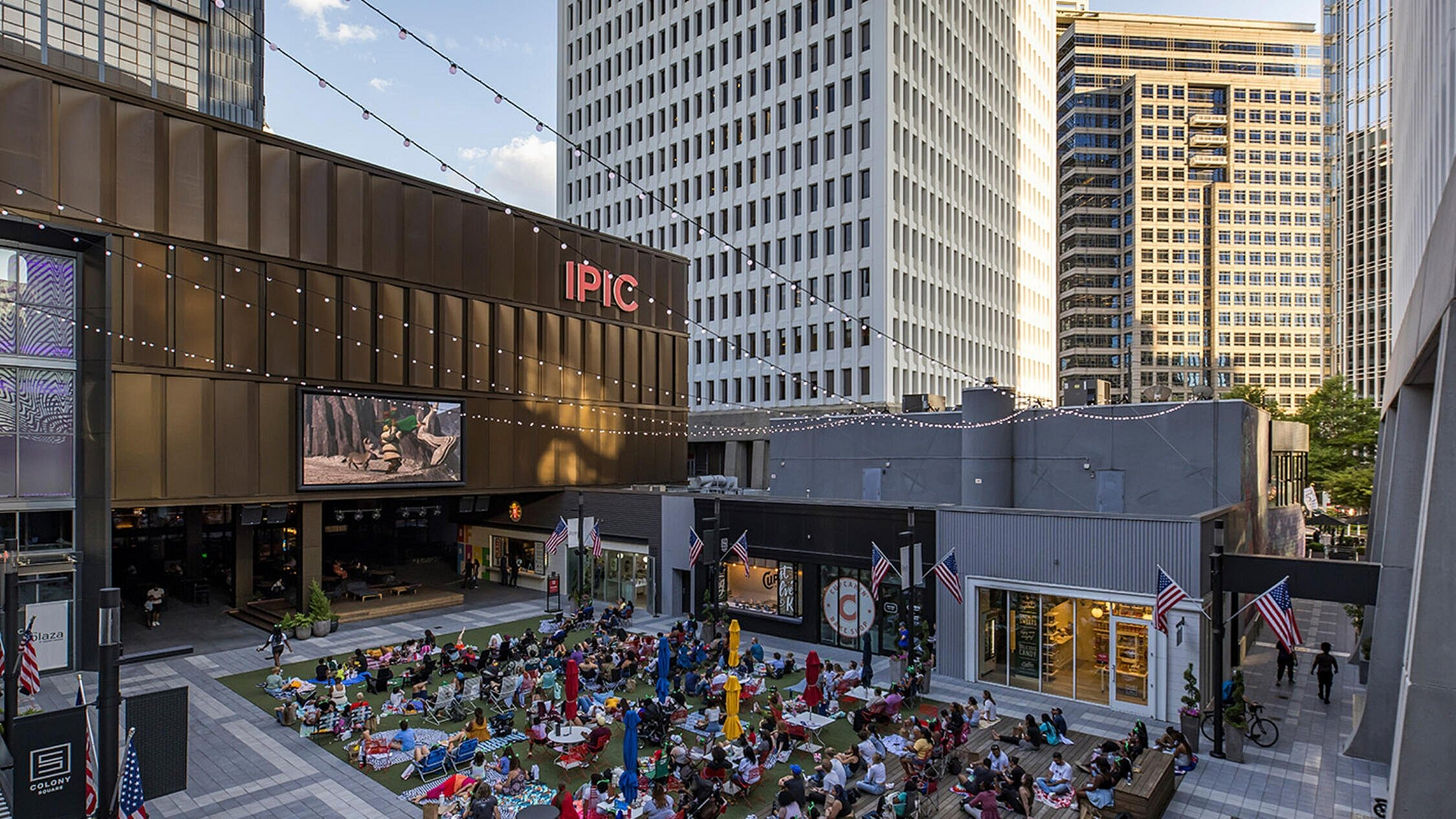 Vibrant urban plaza filled with people enjoying an outdoor movie screening under string lights, surrounded by towering modern buildings.