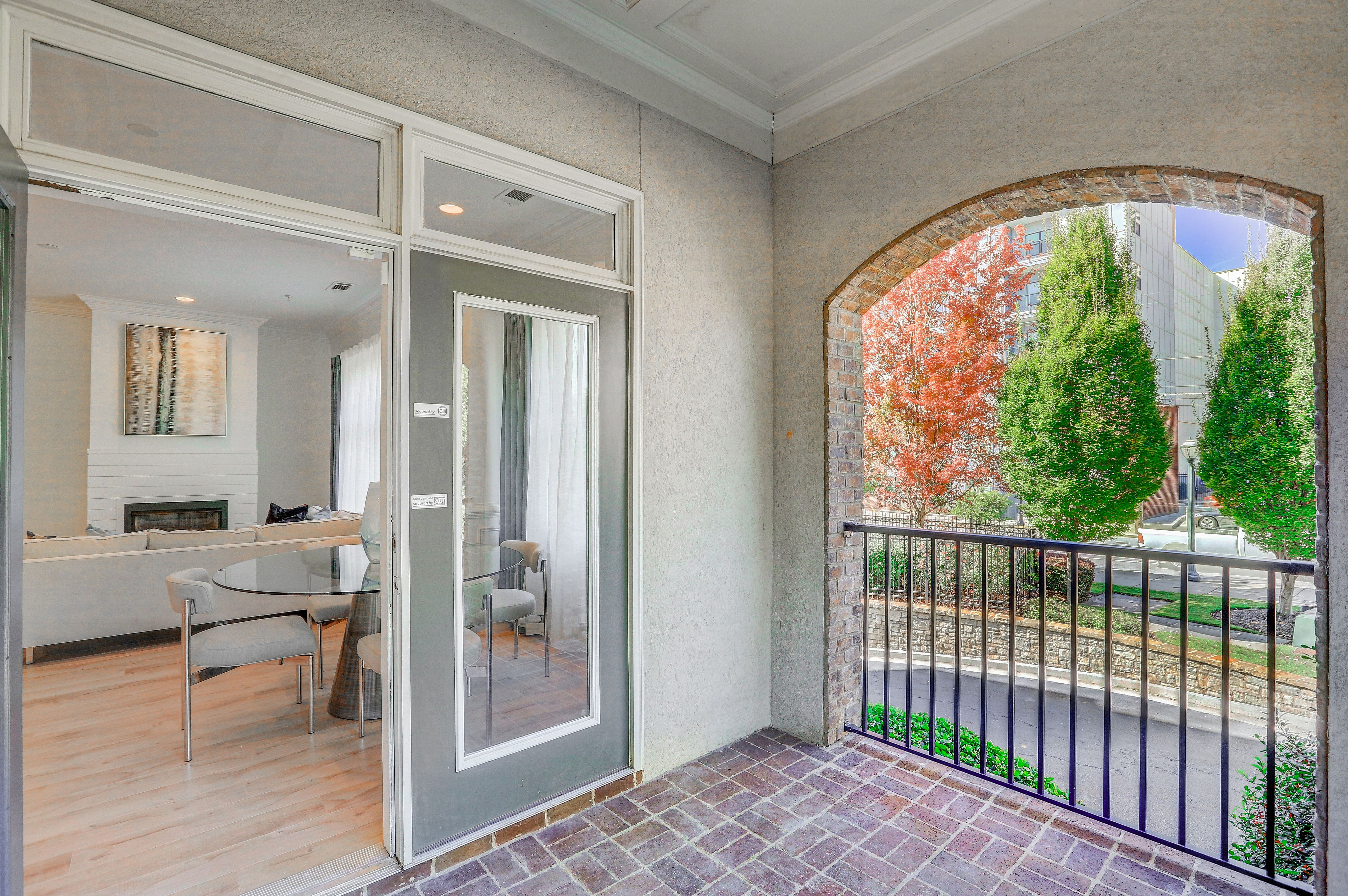 Charming apartment balcony with elegant brickwork, opening into a stylish living area featuring soft seating and a modern glass dining table, surrounded by lush green and autumnal trees.