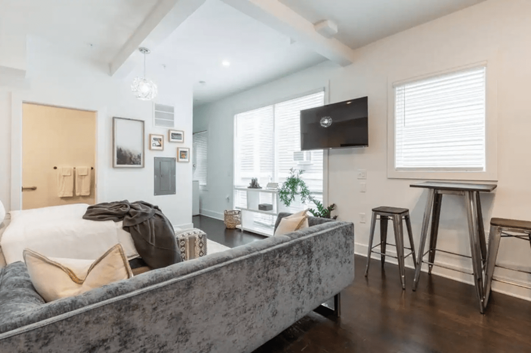 Bright and cozy apartment living space with modern decor, featuring a plush velvet sofa, chic wall art, abundant natural light from large windows, and a sleek mounted TV.