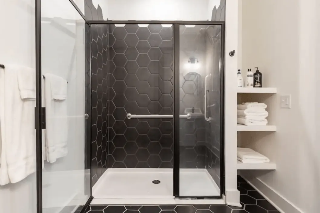 Elegant bathroom with black hexagonal tiles, spacious glass shower, and open shelving for towels.