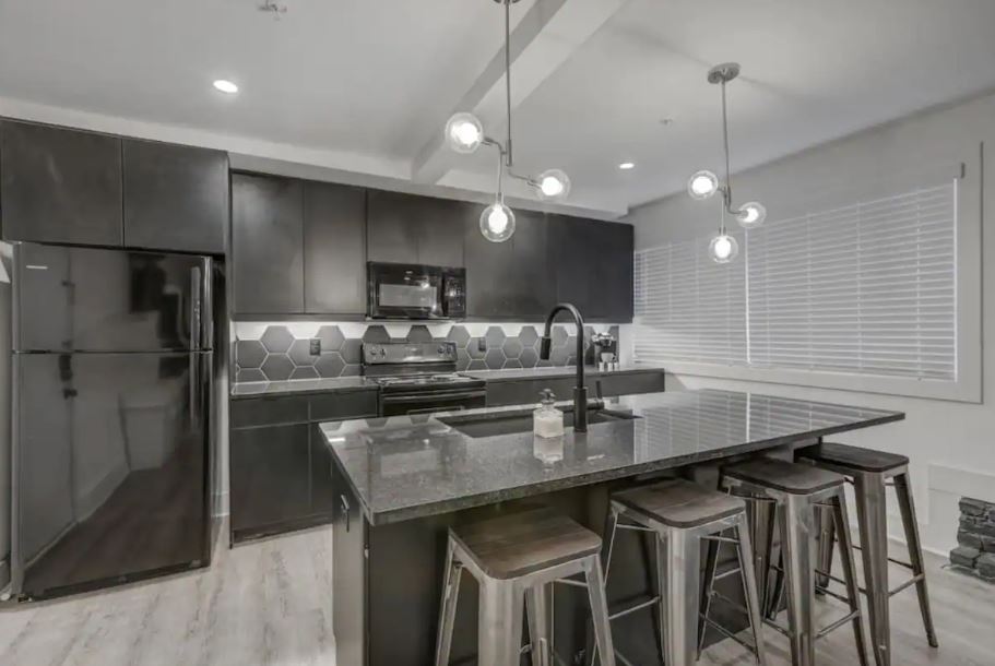 Modern kitchen with sleek black cabinets, a large island with seating, stylish pendant lighting, and stainless steel appliances.