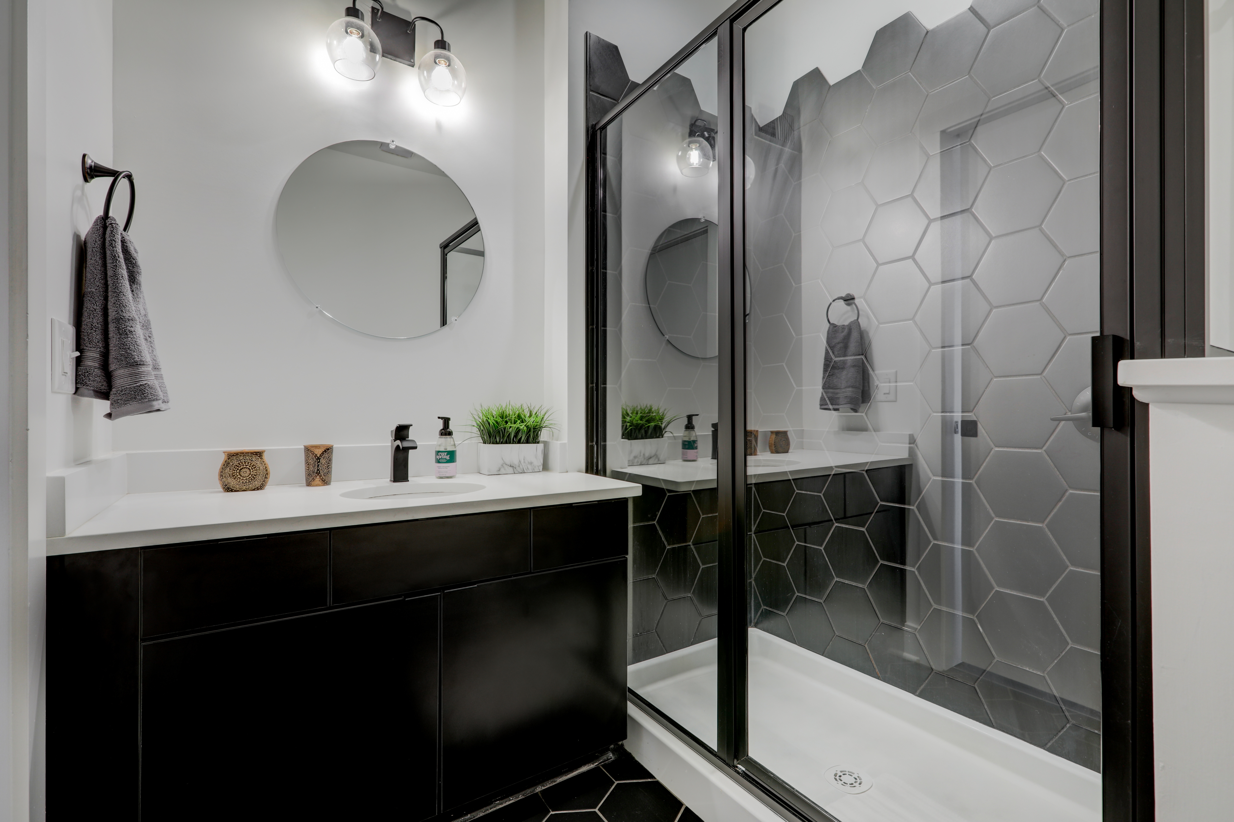 Sleek and modern bathroom featuring a stylish black vanity, round mirror, and glass-enclosed shower with hexagonal tiles.