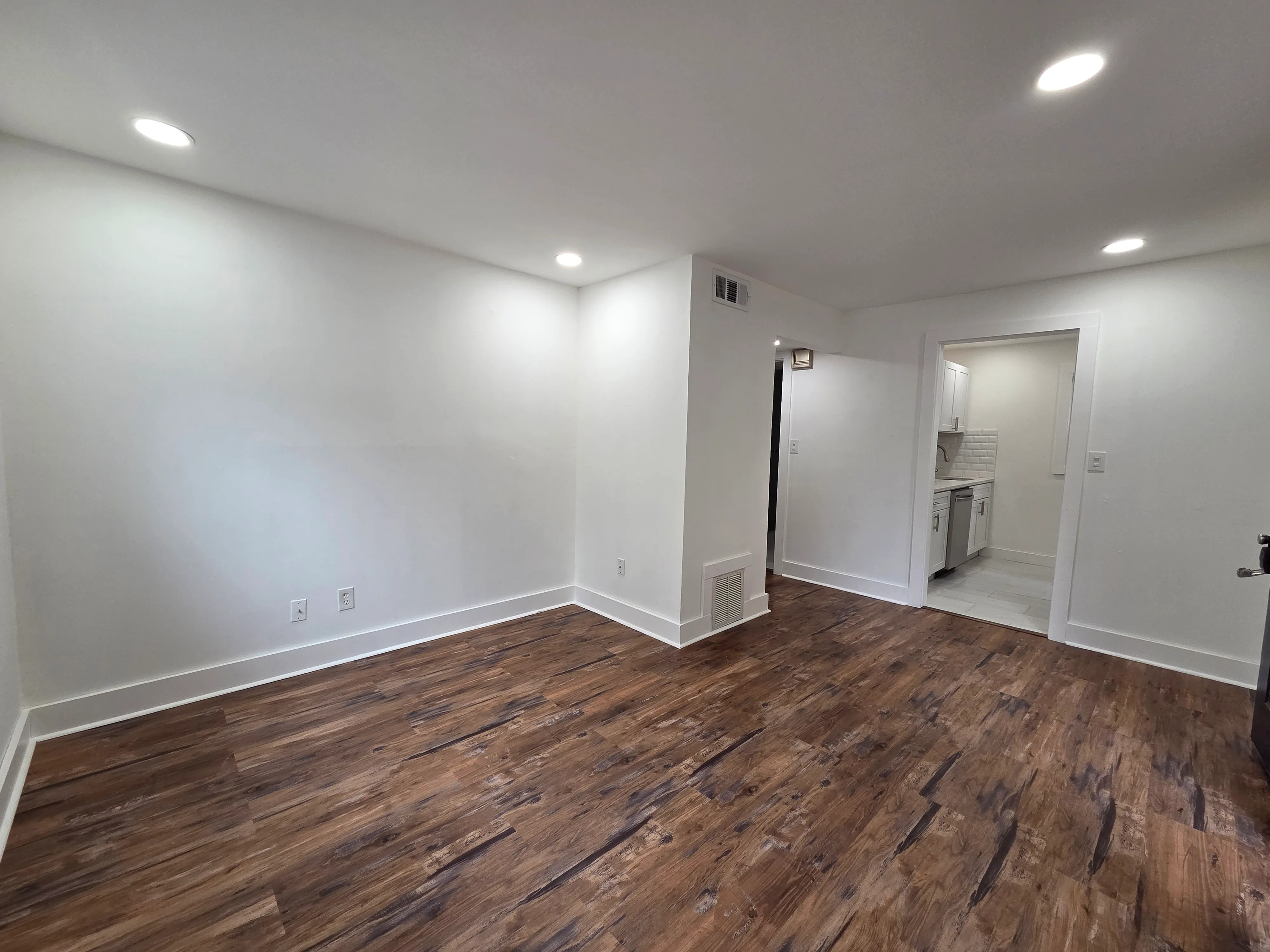 Inviting living space with elegant wood flooring and modern recessed lighting leading to a sleek kitchen area.