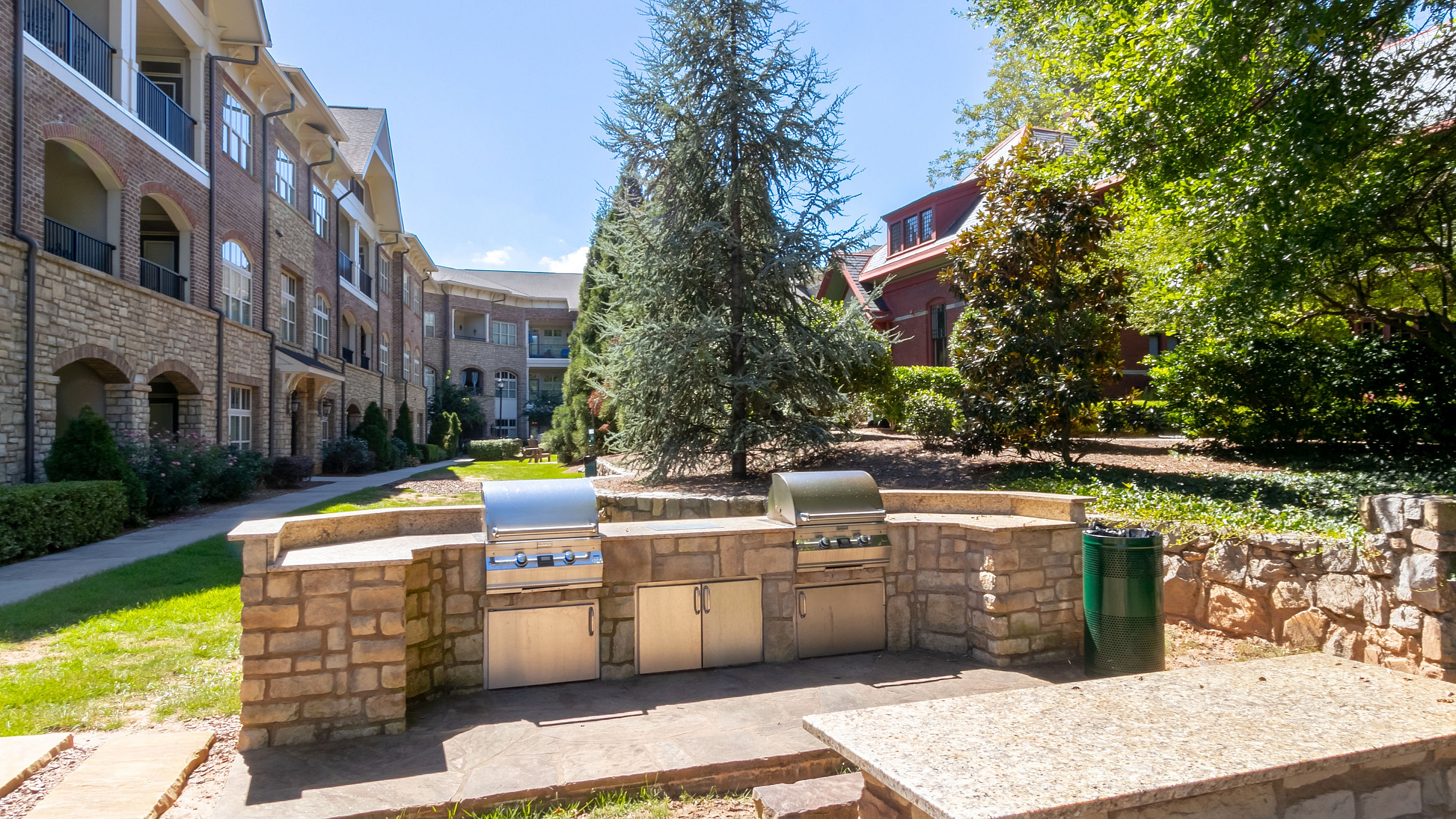 Charming apartment building with elegant stone facade, featuring a well-equipped outdoor grilling area surrounded by lush greenery and trees.