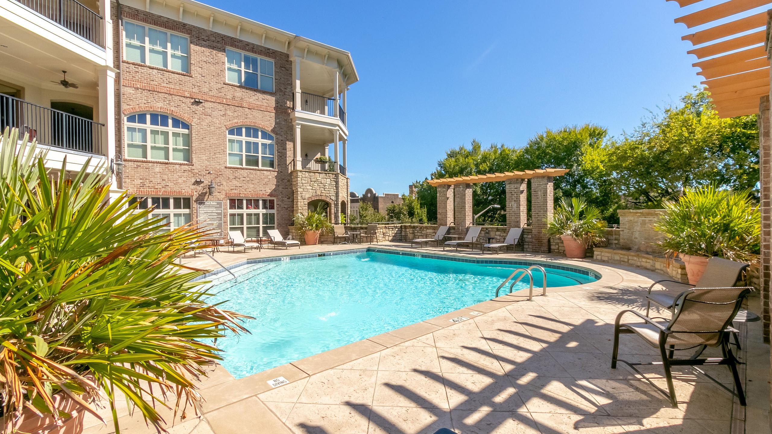 Luxurious apartment complex with a pristine swimming pool, surrounded by elegant lounge chairs and lush greenery under a clear blue sky.