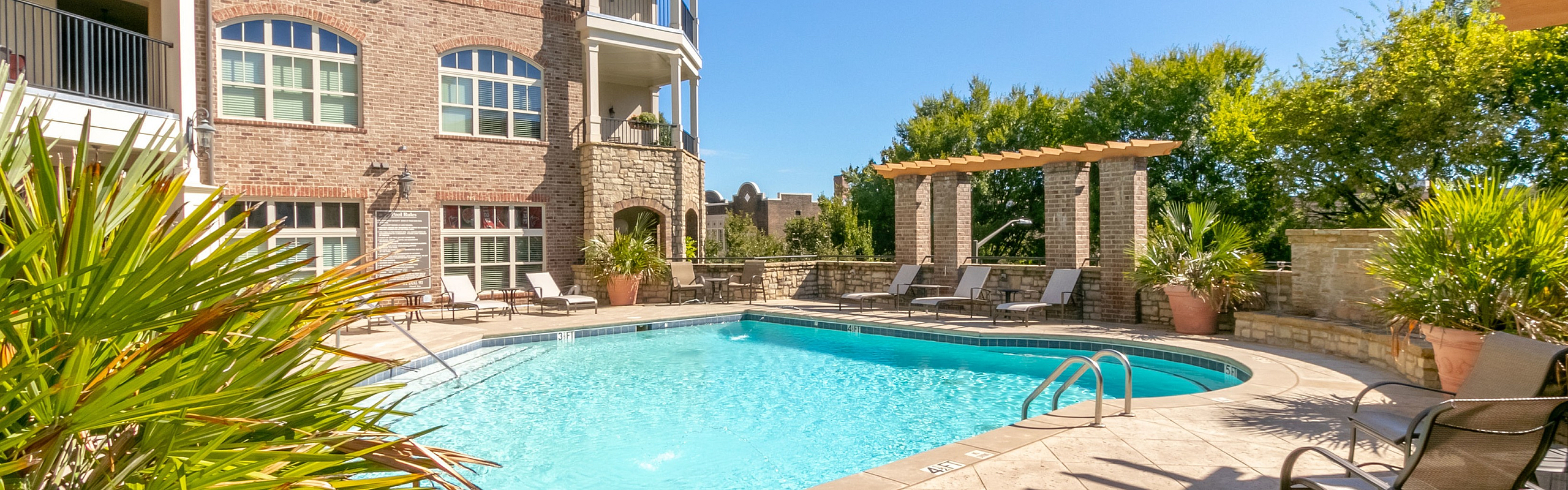 Inviting apartment complex courtyard with a sparkling pool, sun loungers, and lush greenery under a clear blue sky.
