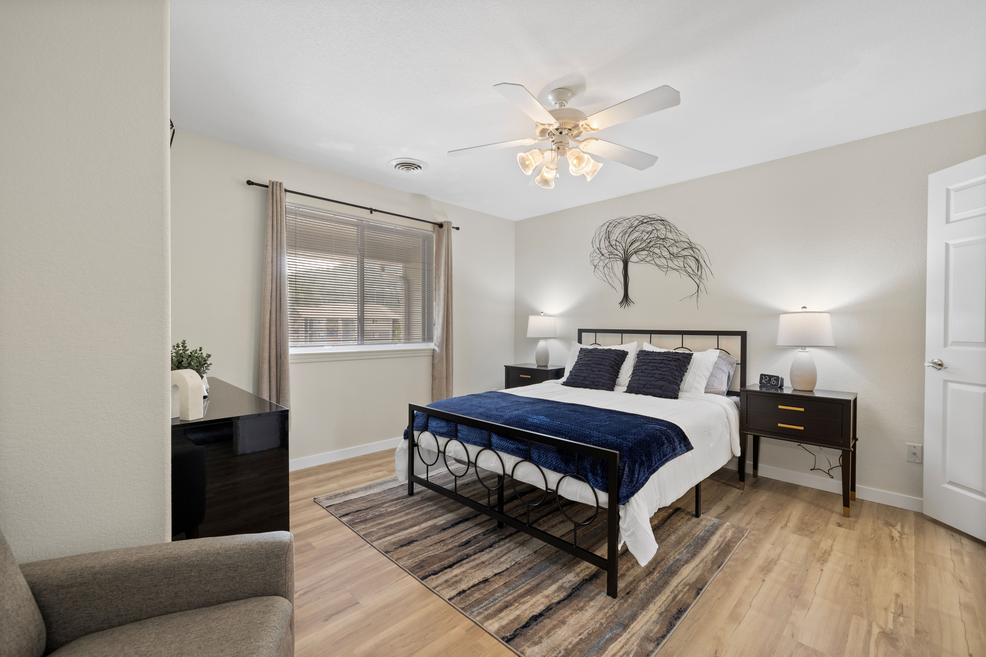 Inviting bedroom featuring a cozy bed with stylish decor and natural light from a large window, highlighted by elegant lighting fixtures and wooden flooring.
