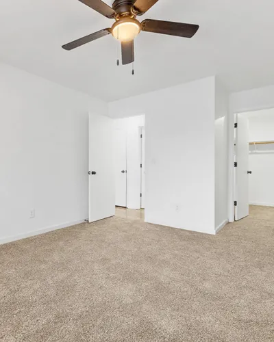Bright and airy bedroom with plush carpeting and a modern ceiling fan, featuring open closet space and minimalist design.