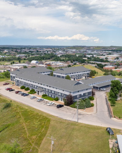 Expansive aerial view of a modern apartment complex surrounded by lush greenery and open fields, offering ample parking and a serene suburban setting.