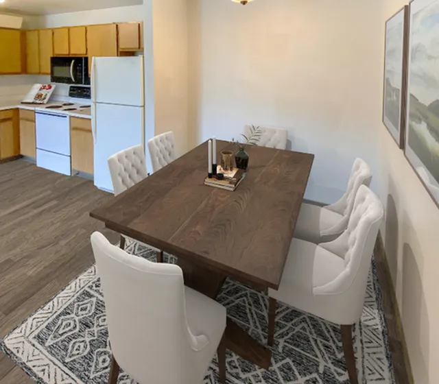 Elegant dining area with a dark wood table and plush white chairs adjacent to a cozy kitchen, featuring modern appliances and warm wood cabinetry.