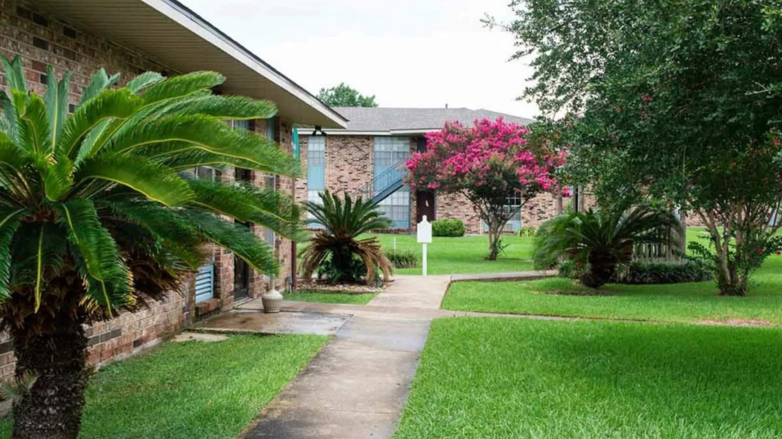 Charming brick apartment complex surrounded by lush greenery and vibrant flowering trees, offering serene living spaces.