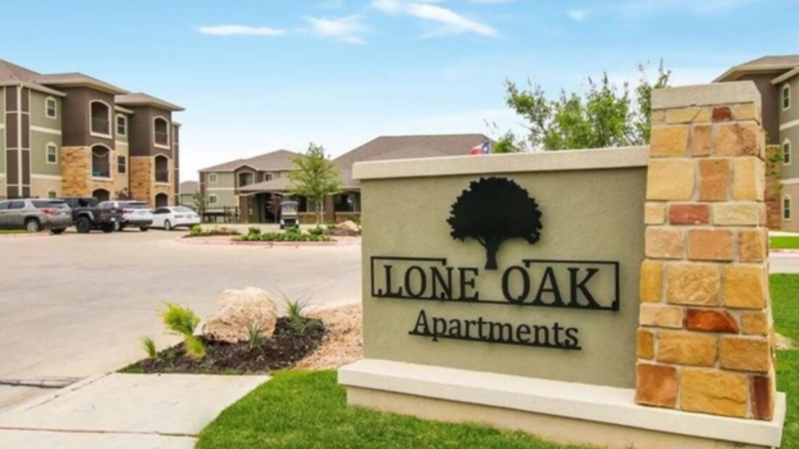 Elegant entrance of Lone Oak Apartments with modern architecture and lush landscaping under a bright blue sky.