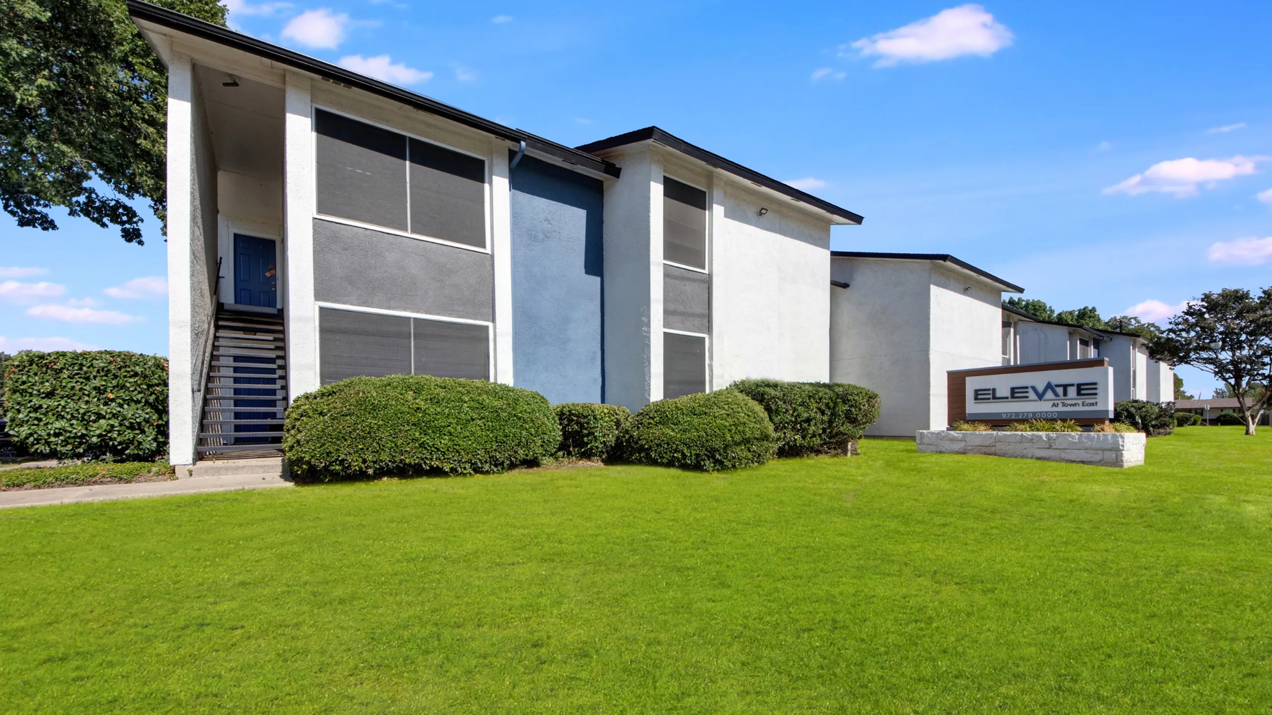 Modern apartment complex with sleek design, lush green lawns, and vibrant shrubs under a bright sky, featuring a welcoming entrance and stylish signage.