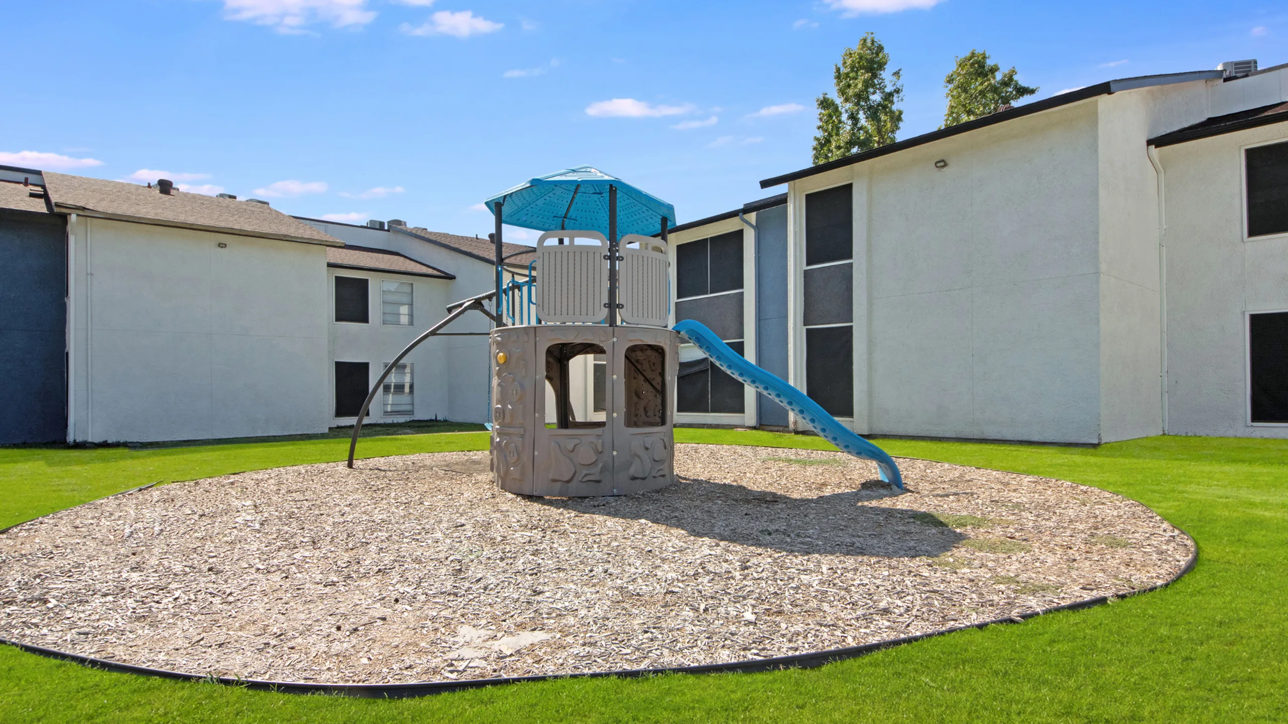 Charming apartment complex with a vibrant playground area and lush green surroundings under a clear blue sky.