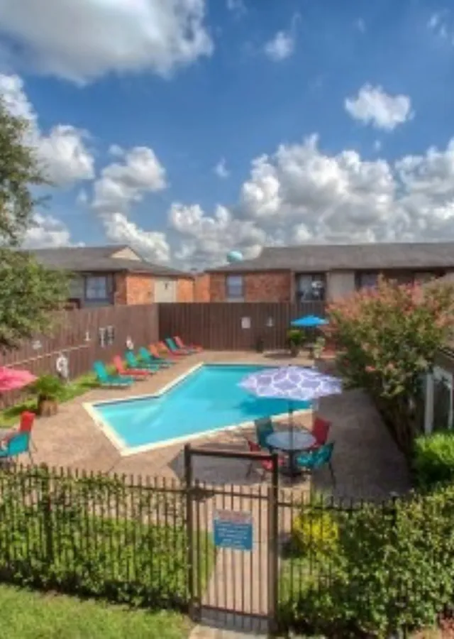 Inviting apartment community pool with colorful lounge chairs and umbrellas, surrounded by lush greenery and clear blue skies.