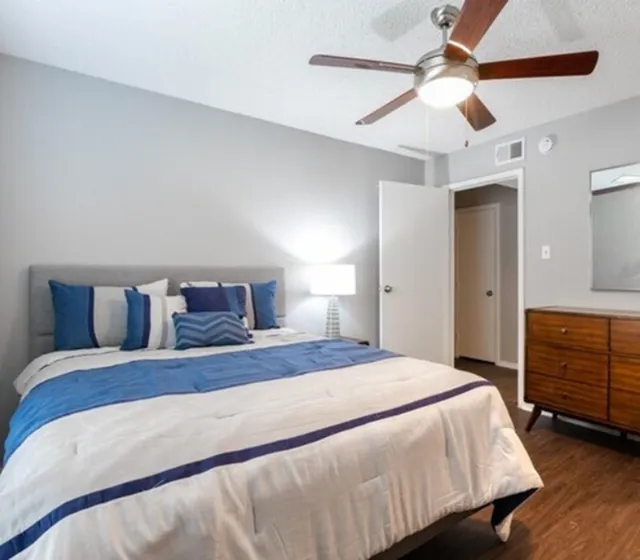 Inviting bedroom with modern decor, plush bedding in blue and white, warm wooden dresser, and ceiling fan for comfort.