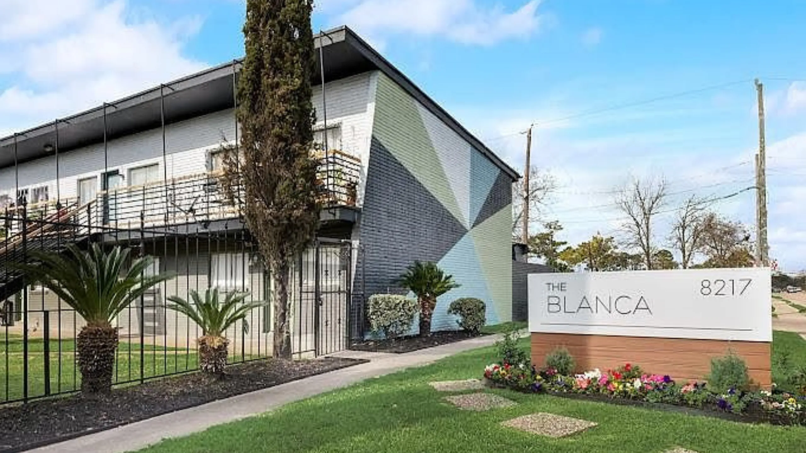 Modern apartment exterior of The Blanca featuring geometric wall design, lush greenery, and a welcoming path.