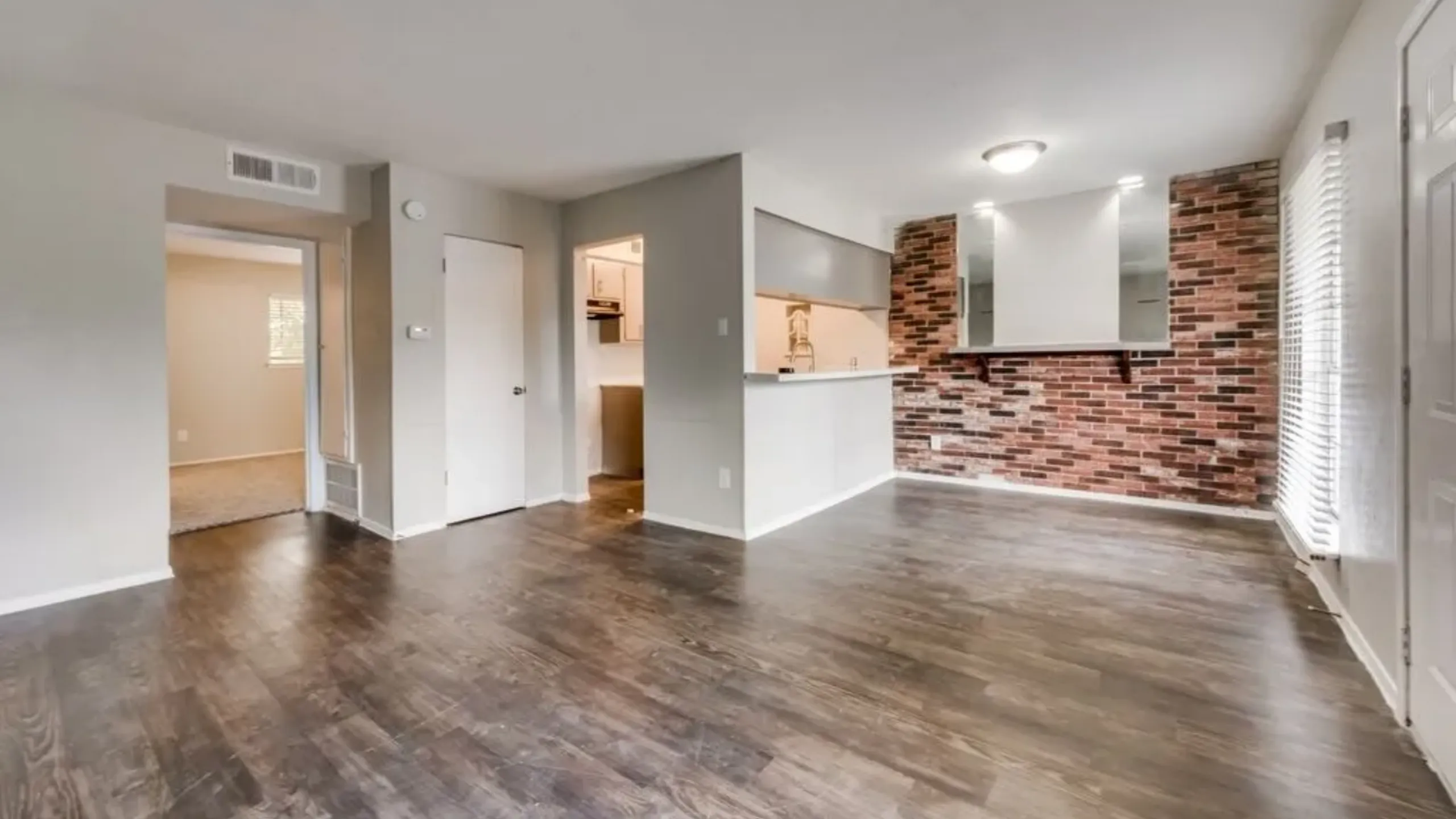 Modern apartment interior with sleek wood flooring, an open kitchen with brick accent wall, and ample natural light.