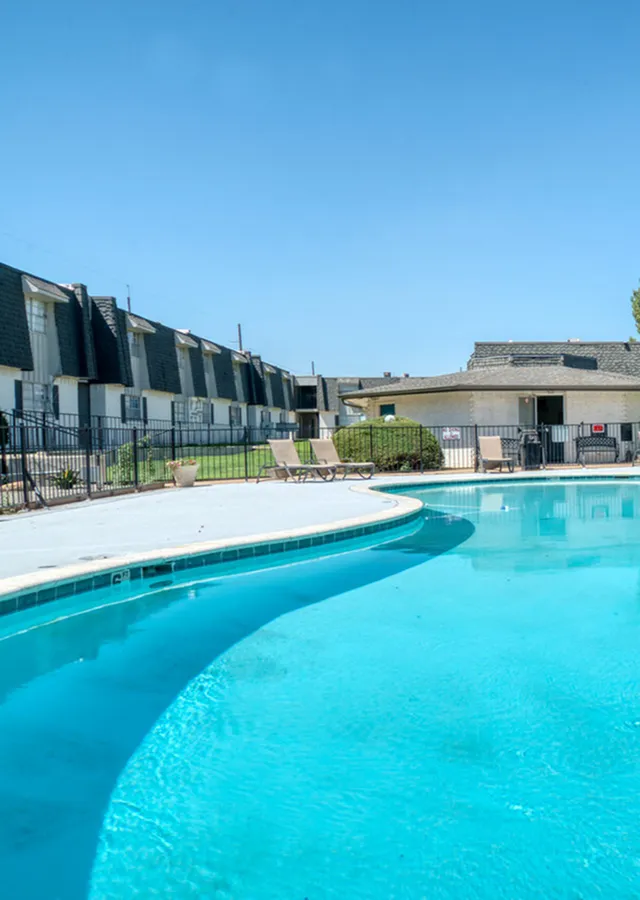 Inviting apartment complex with a pristine swimming pool, surrounded by modern townhouses and lush greenery under a clear blue sky.