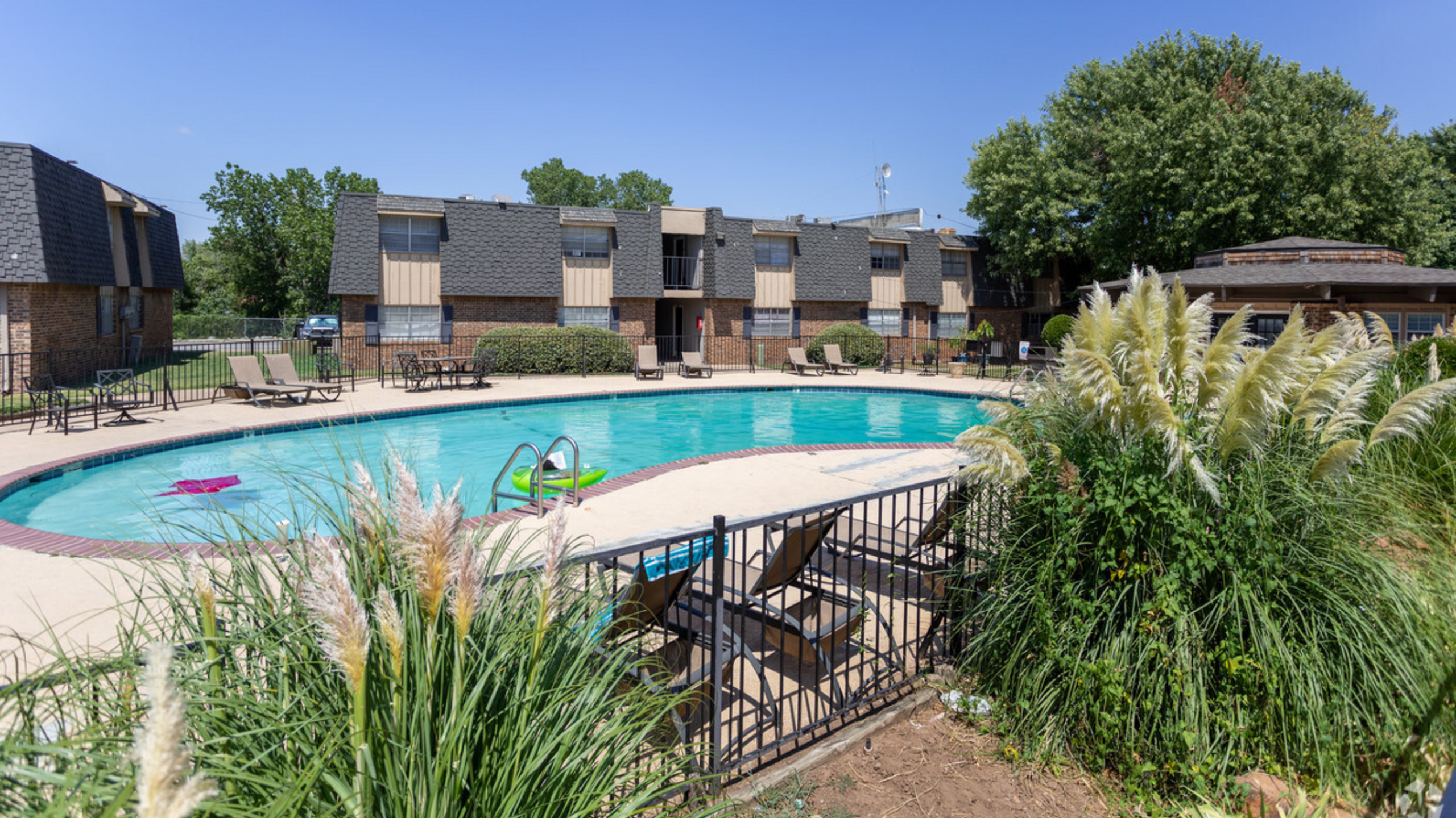 Inviting apartment complex with a sparkling pool, surrounded by lush greenery and cozy loungers under a clear blue sky.