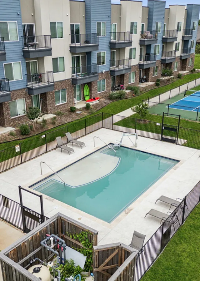 Contemporary apartment complex with balconies overlooking a pristine outdoor pool, surrounded by lush greenery and a nearby sports court.