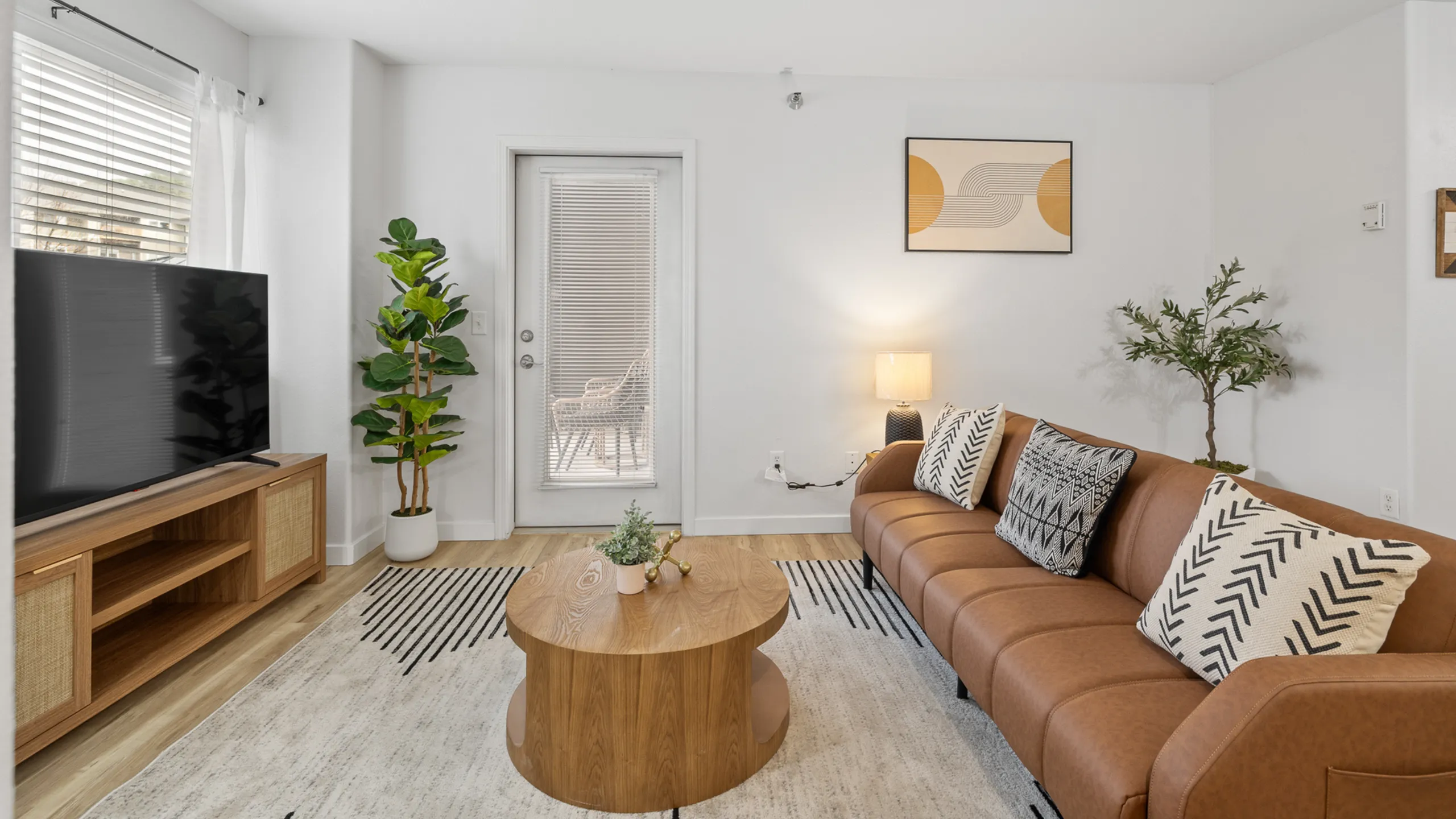 Inviting living room with a sleek brown leather sofa, stylish cushions, and warm lighting. Features a large TV, vibrant indoor plants, and chic decor, offering a perfect blend of comfort and modern elegance.