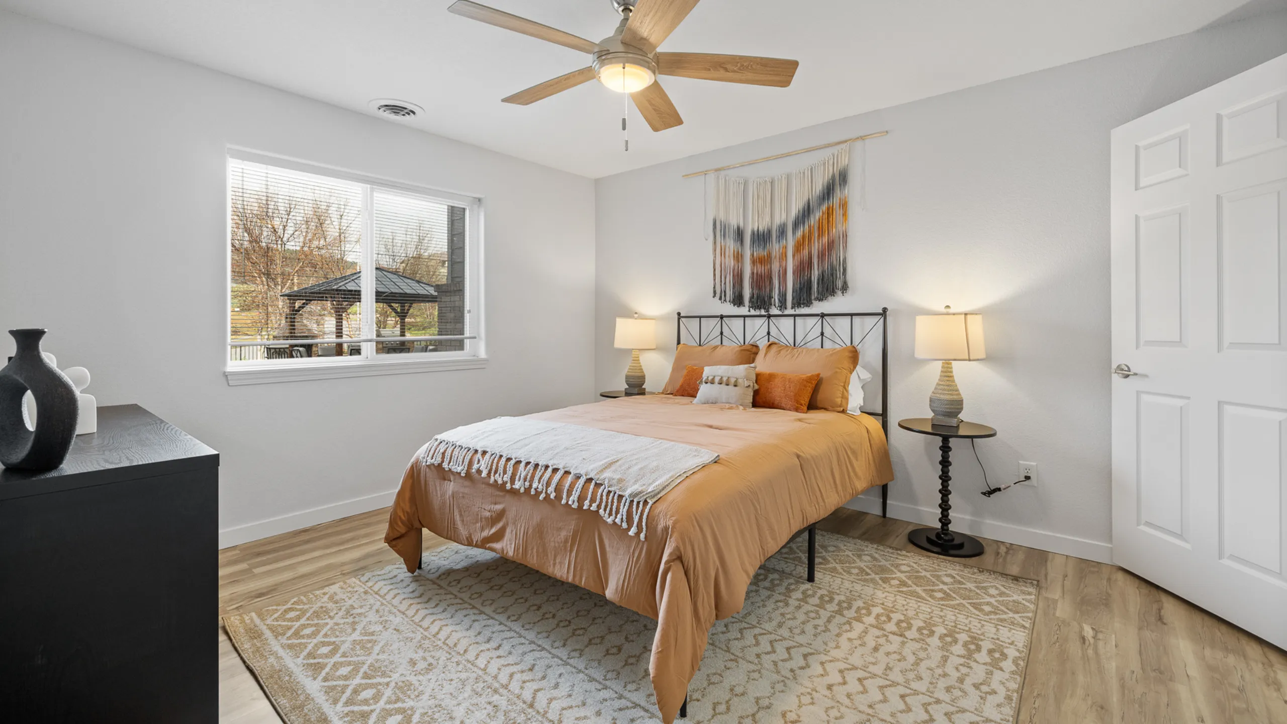 Inviting bedroom with elegant decor, featuring a cozy bed with warm-toned bedding, stylish wall art, and a scenic window view. Modern ceiling fan and chic side lamps enhance the comfort and aesthetics.