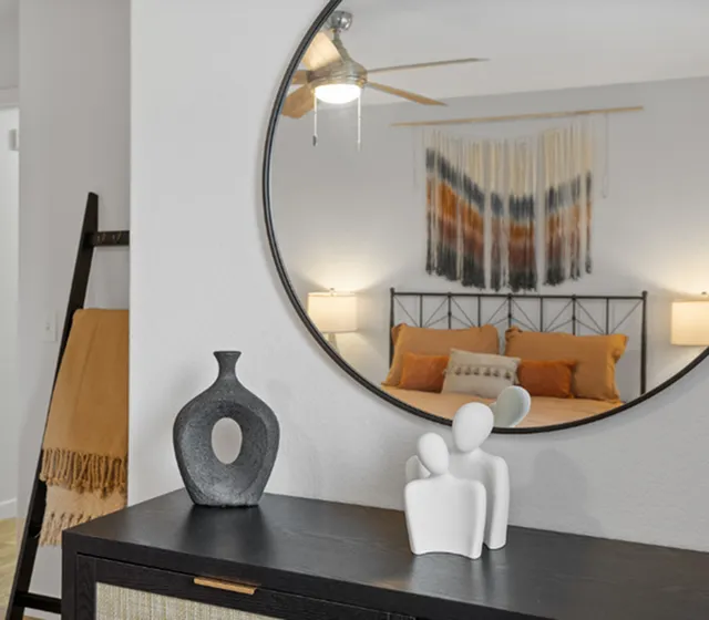Modern bedroom with cozy decor reflected in a round mirror, featuring a stylish bed with warm-toned pillows and artistic wall hanging.