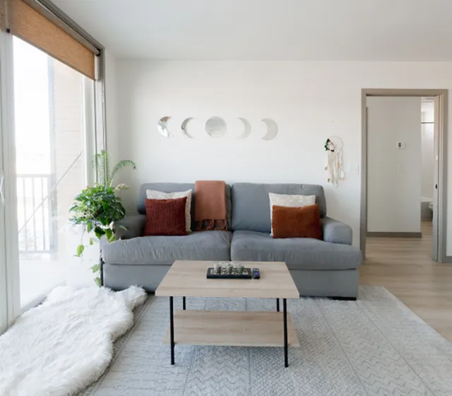 Bright and inviting living room with a cozy gray sofa, accent pillows, and a stylish coffee table. Features large windows, lush greenery, and decorative wall art for a serene ambiance.