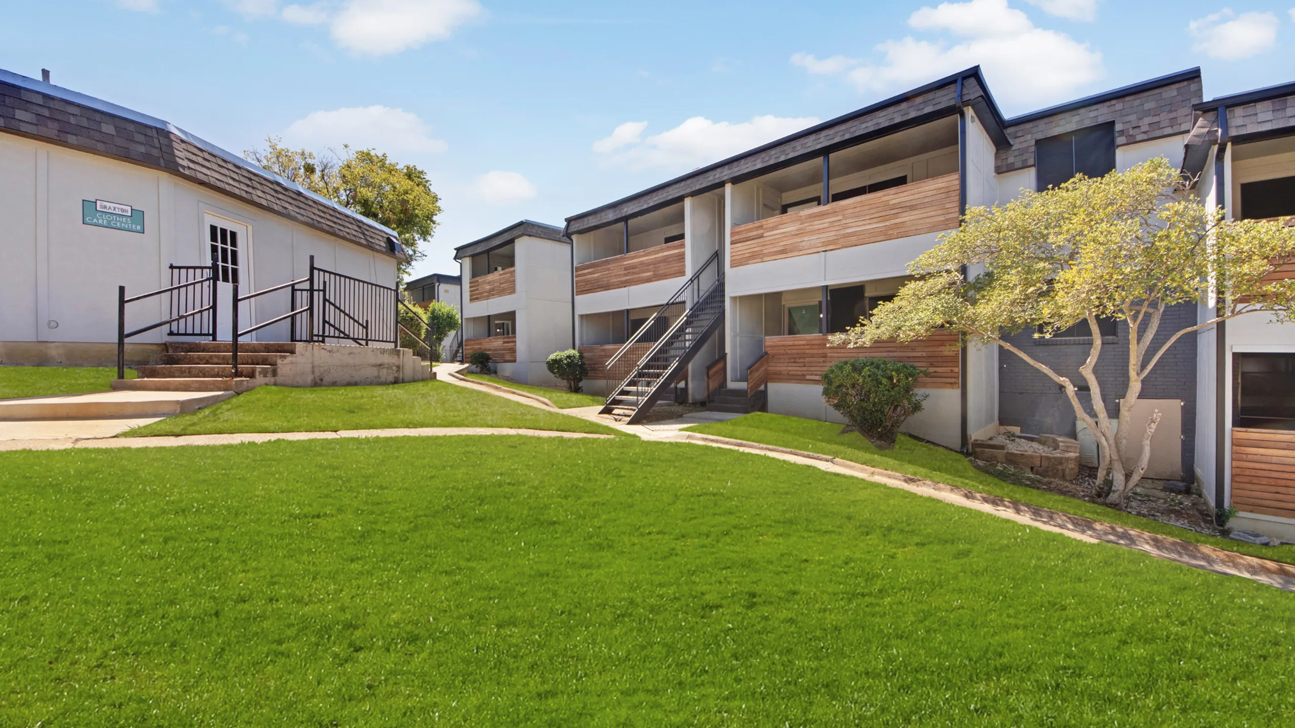 Charming apartment complex with modern design, featuring wooden balcony accents, lush green lawn, and sunny pathways.