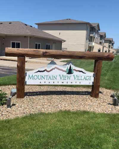 Welcome to Mountain View Village Apartments, showcasing a rustic wooden sign with a serene backdrop of multi-unit buildings under a clear blue sky.