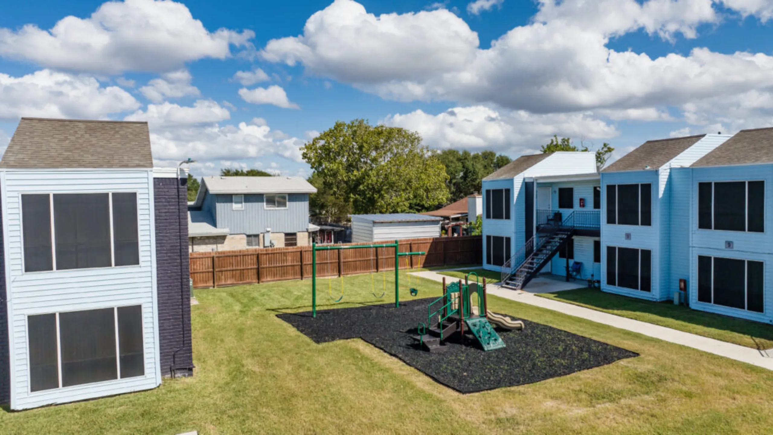 Charming apartment building surrounded by lush greenery and playful kids' playground under a clear blue sky.
