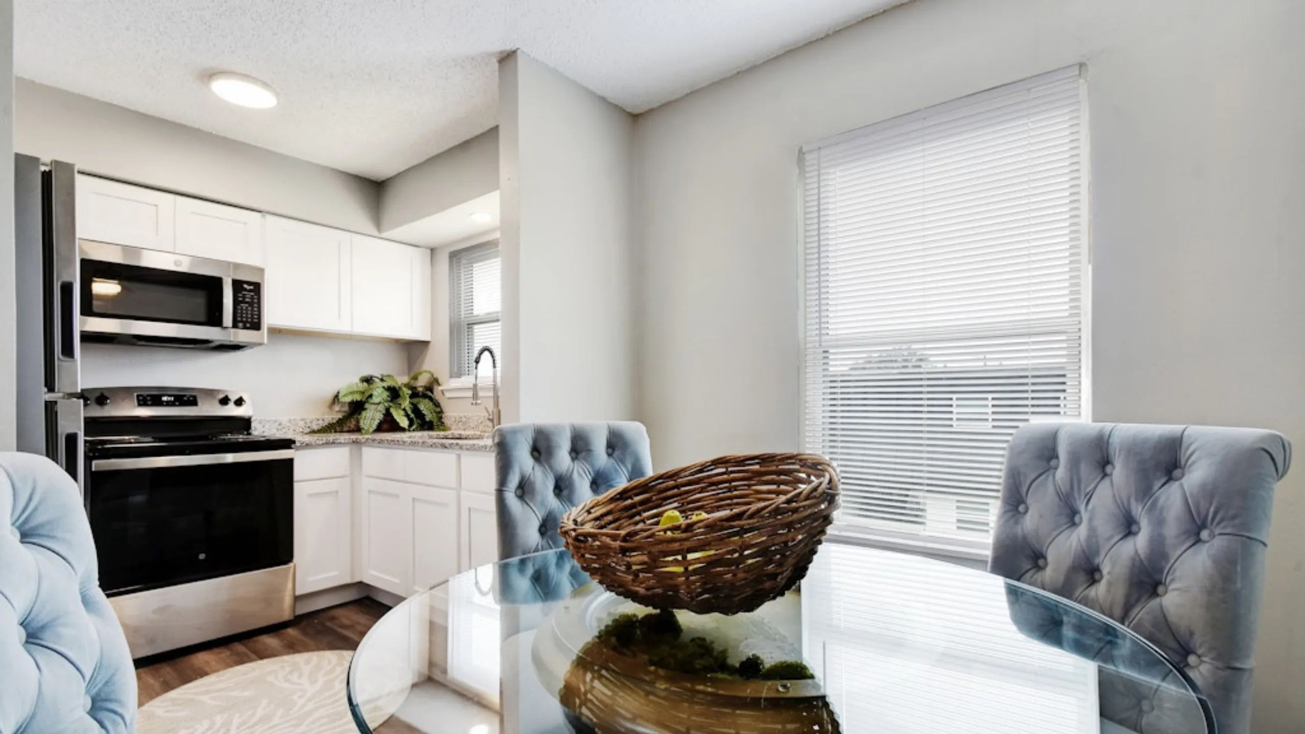 Modern kitchen and dining area with stainless steel appliances, elegant glass table, and plush chairs, bathed in natural light.