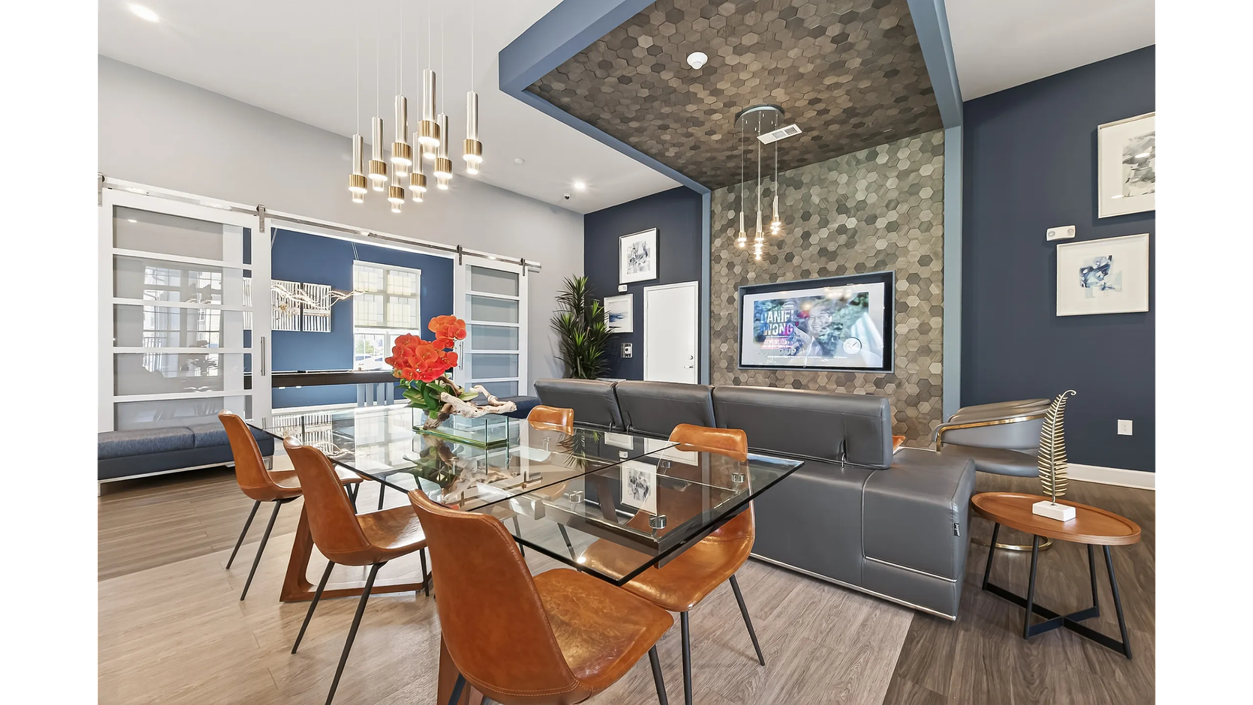 Modern living area featuring a sleek glass dining table, cozy leather chairs, and an elegant light fixture, complemented by stylish wall decor and vibrant red flowers.
