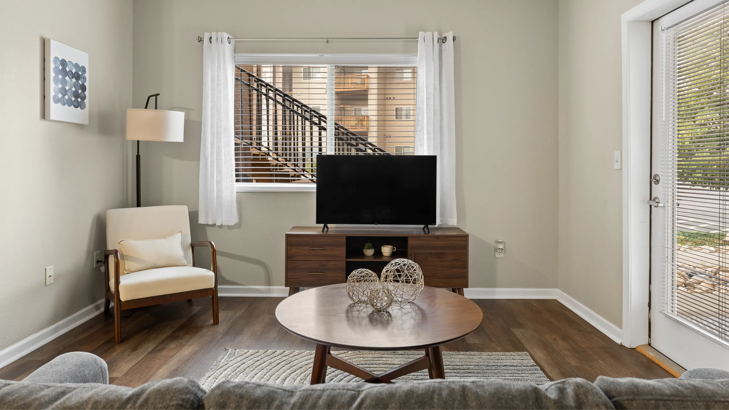 Elegant living room with modern furniture, featuring a cozy armchair, stylish coffee table, and entertainment center with a flat-screen TV, bathed in natural light from large windows.