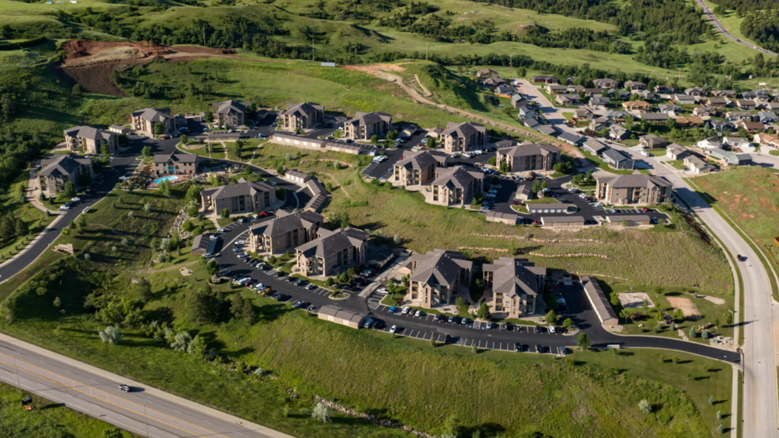 Aerial view of a picturesque residential development nestled in lush greenery, showcasing modern apartment buildings with ample parking and scenic surroundings.