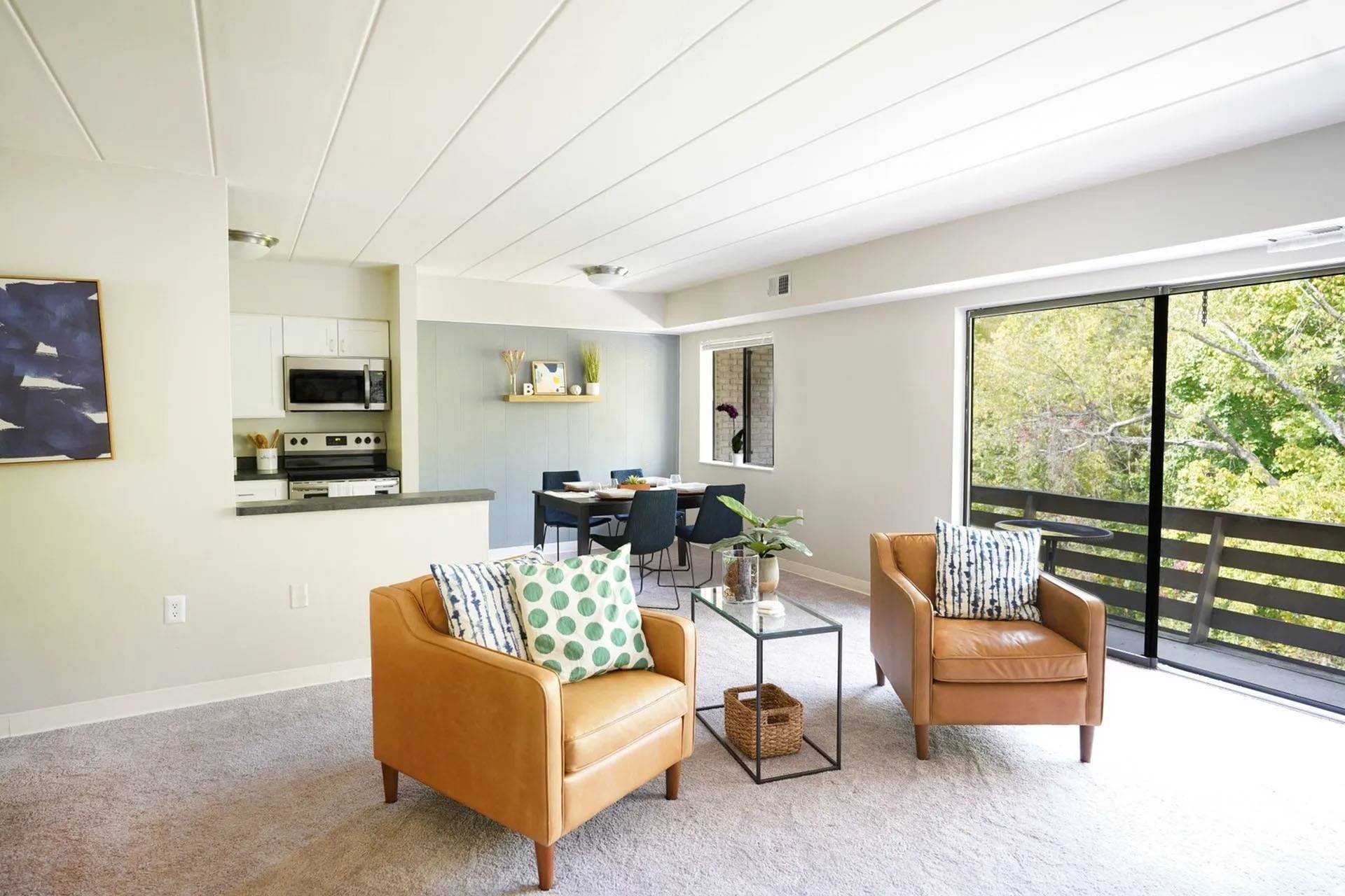 Apartments Near South Park in Bethel Park, PA - Lindenbrooke - Living Room with Carpeted Flooring, Brown Leather Armchairs, Side Table, View of Dining Area and Kitchen, and Sliding Glass Door with Access to Balcony.