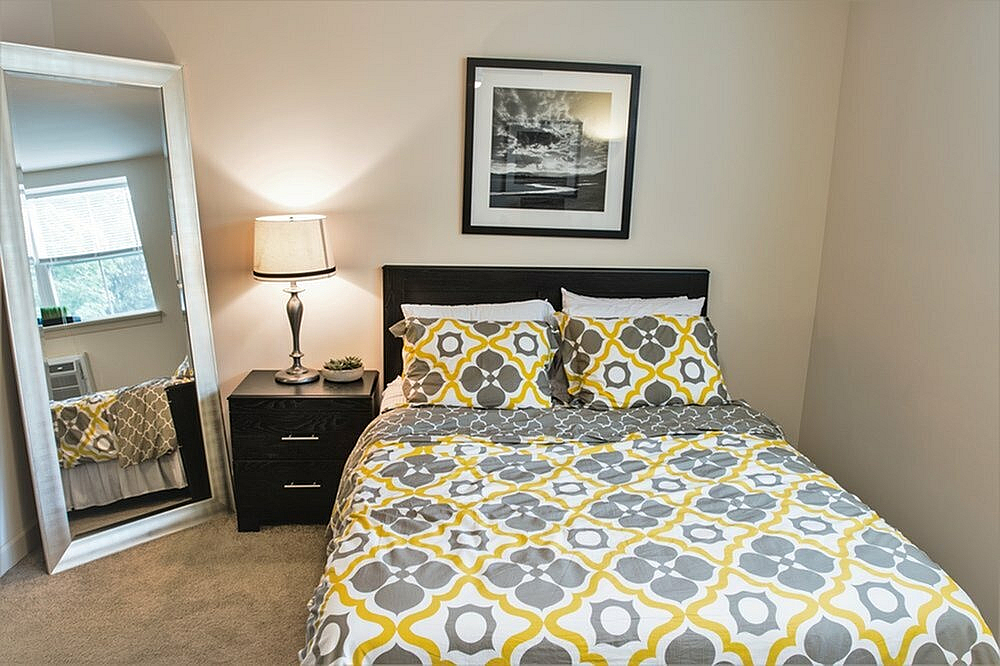 Cozy bedroom featuring a stylish bed with yellow and gray patterned bedding, a nightstand with a lamp, and a large mirror.