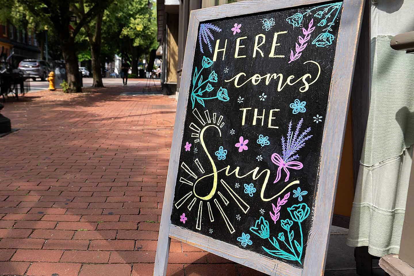 Charming sidewalk chalkboard with colorful floral designs and uplifting 'Here Comes the Sun' message.